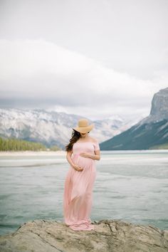 a pregnant woman in a pink dress and hat standing on a rock by the water