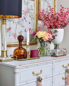 a white dresser topped with lots of flowers next to a wall mounted mirror and lamp