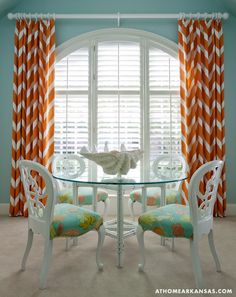 an orange and white dining room with curtains on the window sill, chairs around a glass table