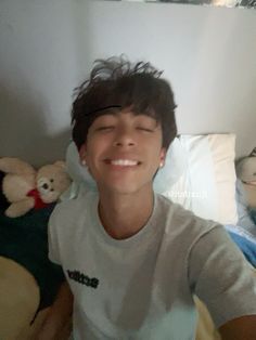 a young man is smiling while sitting on a bed with his eyes closed and stuffed animals in the background