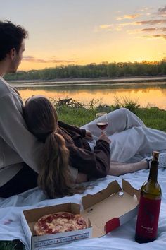 a man and woman sitting on a blanket with pizza and wine in front of them