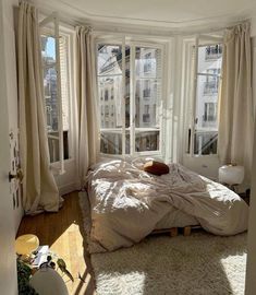 an unmade bed sitting in the corner of a room next to two large windows