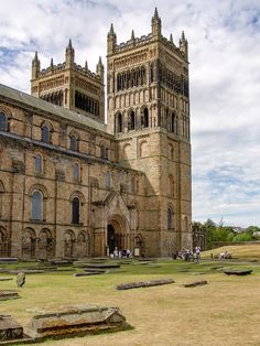 an old building with two towers and people walking around it in the grass near benches