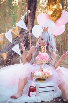 a woman sitting on top of a table with a cake in front of her and balloons behind her