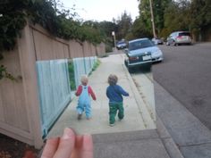 two young boys walking down the sidewalk in front of a fence and cars parked on the side walk