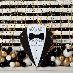 a black and white striped backdrop with gold and silver balloons, tuxedo bow tie sign, and confetti garland