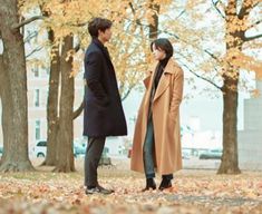 a man and woman standing in front of trees with leaves on the ground looking at each other