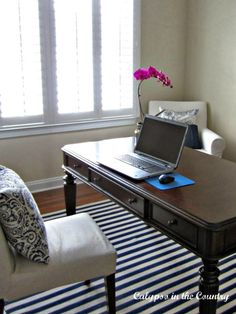 a laptop computer sitting on top of a wooden desk next to a white chair and window