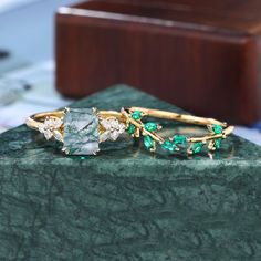 two rings with green and white stones sitting on top of a marble slab next to a wooden box