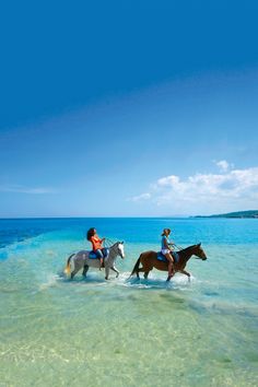 two people riding horses through shallow water on the ocean's edge, with blue sky in the background