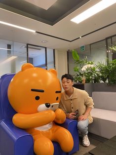 a man sitting next to a giant teddy bear in an office setting with plants behind him