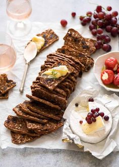 a table topped with bread, fruit and crackers next to two glasses of wine