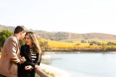 a man and woman standing next to each other near a body of water with hills in the background