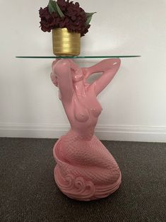 a pink mermaid figurine sitting on top of a glass table next to a vase with flowers