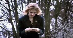 a woman standing in the woods looking at her cell phone while wearing a black coat