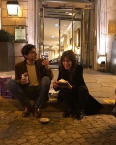 two people sitting on the ground eating food and drinking coffee in front of a building