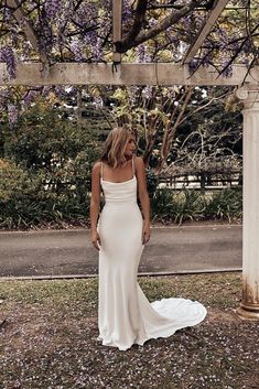 a woman in a white wedding dress standing under a pergolia covered arbor with purple flowers