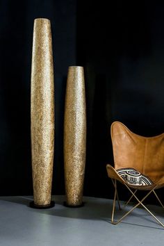 two tall gold vases sitting next to each other on a gray floor in front of a chair