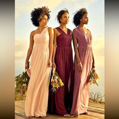 three women in long dresses standing on a wooden platform near the ocean and holding flowers