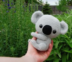 a hand holding a small crocheted koala bear in front of some bushes