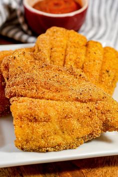 some fried food on a white plate next to a bowl of sauce and a fork