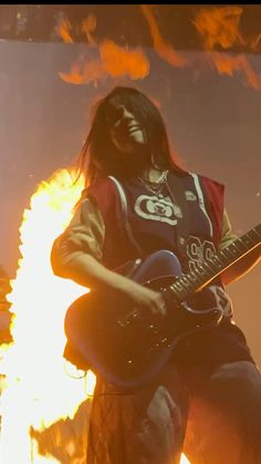 a man with long hair playing an electric guitar in front of a large fire filled background