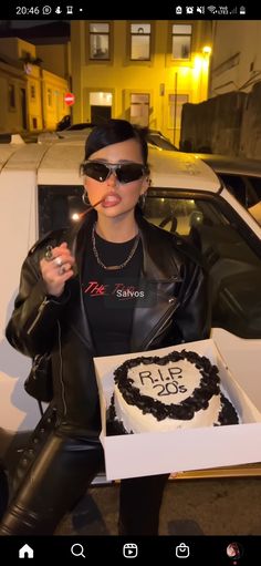 a woman in black jacket and sunglasses holding up a box with a cake on it