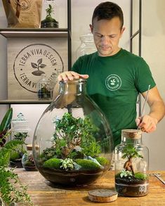 a man is working with plants in a large glass vase on a table next to other items