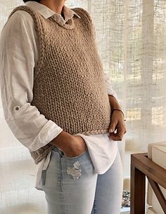 a woman standing in front of a window wearing a knitted sweater vest and jeans