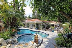 an outdoor swimming pool surrounded by trees and rocks in front of a house with a covered patio