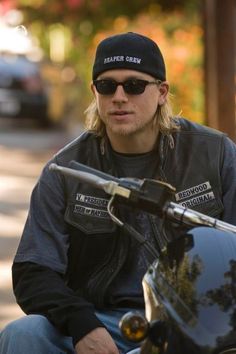 a man wearing sunglasses and a leather jacket sits on a motorcycle with his hat on