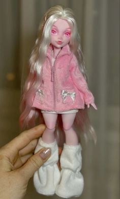 a hand is holding a doll with long blonde hair and pink coat on it's head