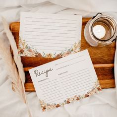 two recipe cards sitting on top of a wooden tray next to a candle and some feathers