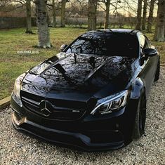 the front end of a black mercedes cla parked on gravel road next to trees