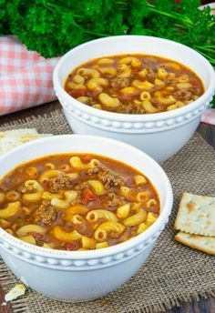 two white bowls filled with pasta and meat soup on top of a table next to crackers