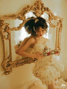 a woman in a white dress is holding a tea cup and looking at herself in the mirror