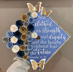 a blue graduation cap decorated with paper flowers and butterfly decorations is held up by a person's hand