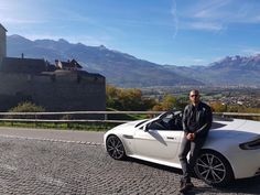 a man standing next to a white sports car in front of a castle with mountains in the background