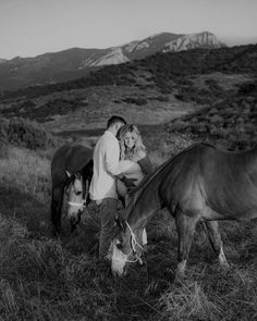 a man and woman are standing next to two horses in the grass with mountains in the background