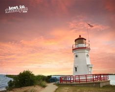 a white lighthouse with a red railing near the water at sunset or sunrise, and an american flag flying in the background