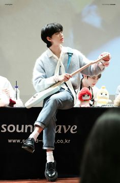 a man sitting on top of a table with a guitar in his hand and stuffed animals behind him