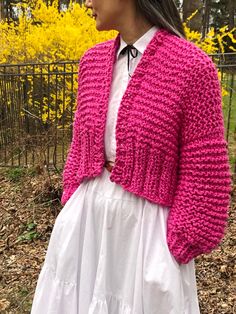 a woman wearing a pink knitted cardigan and white dress standing in front of yellow flowers