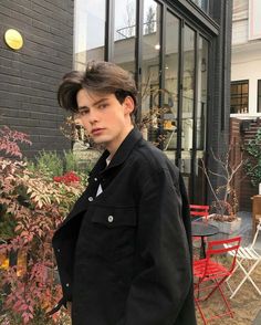 a young man standing in front of a black brick building with red chairs and tables