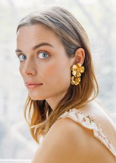 a woman with blue eyes is wearing gold flower earrings in front of a window