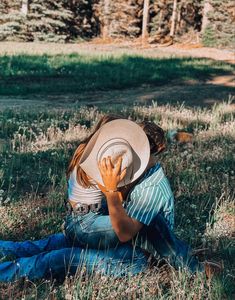 a person sitting in the grass with a hat on their head and one hand up to his face