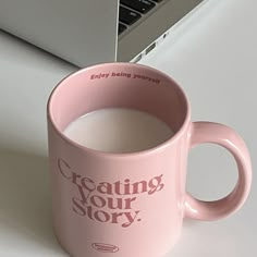 a pink coffee mug sitting on top of a white table next to a laptop computer