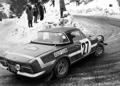 an old race car driving down a snow covered road with people standing in the background