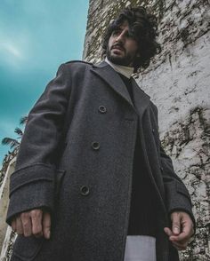 a man standing in front of a stone wall wearing a long coat and white shirt