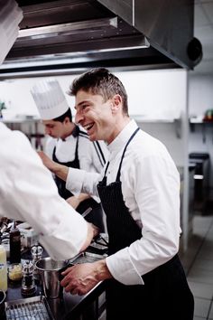 two chefs are smiling while preparing food in a commercial kitchen, one is wearing an apron and the other is holding a knife