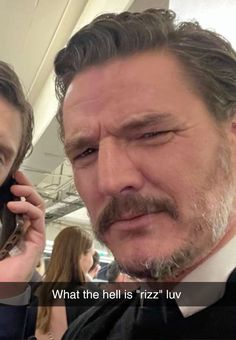 a man with a mustache talking on a cell phone next to another man in a suit and tie